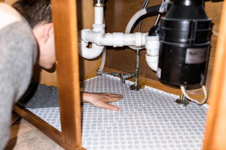 A plumber holding a wrench and inspecting a new residential piping system under a kitchen sink